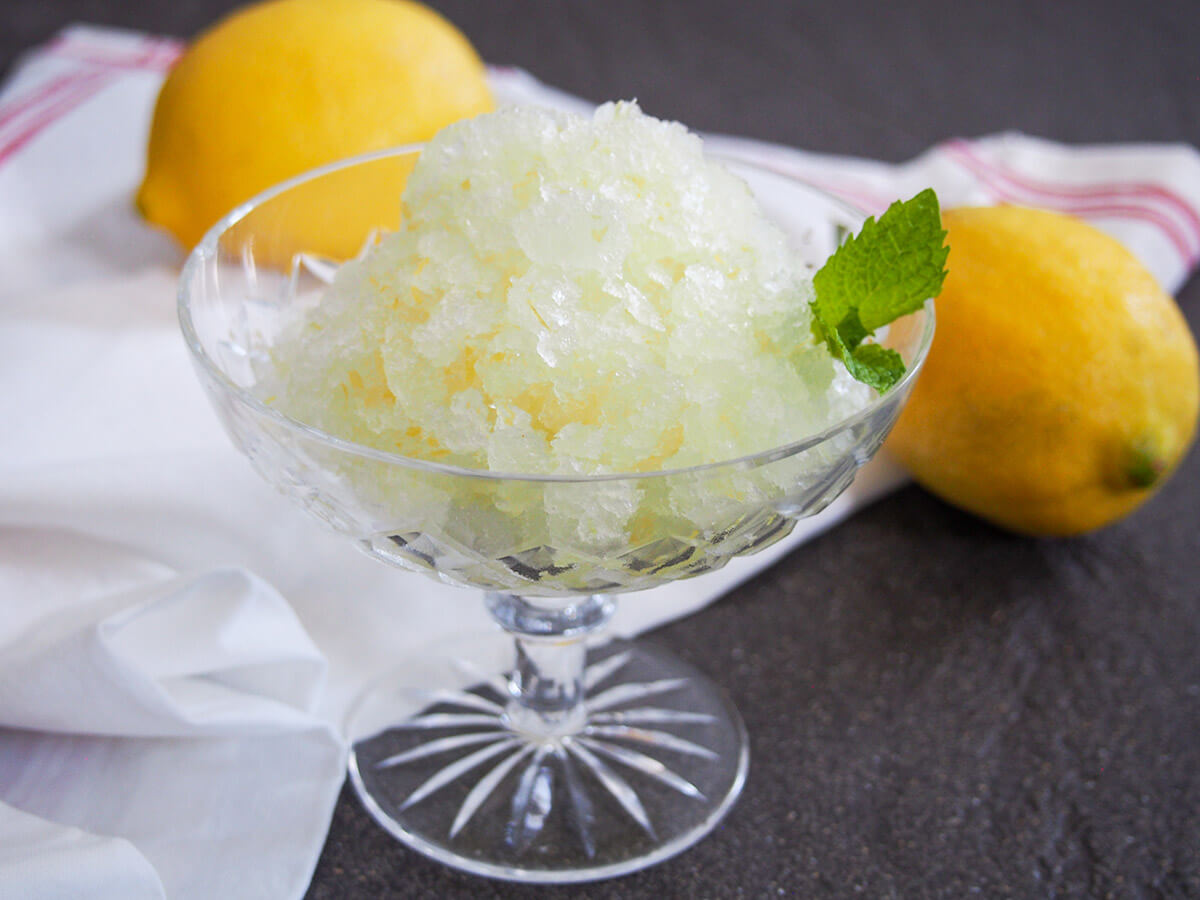 On a warm day, lemon granita is a wonderfully refreshing treat that's perfect to cool you down. It easy to make with a great sweet-tart balance. You'll want it on standby all summer long. glass of lemon granita with two lemons behind glass