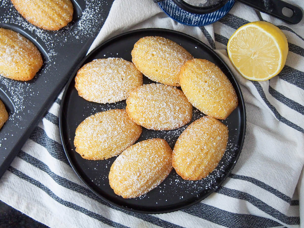 Lemon madeleines are a classic French treat. This small batch recipe makes the perfect amount to enjoy the light, sweet and gently crisp deliciousness at its best. plate of lemon madeleines with half lemon to one side and pan to other