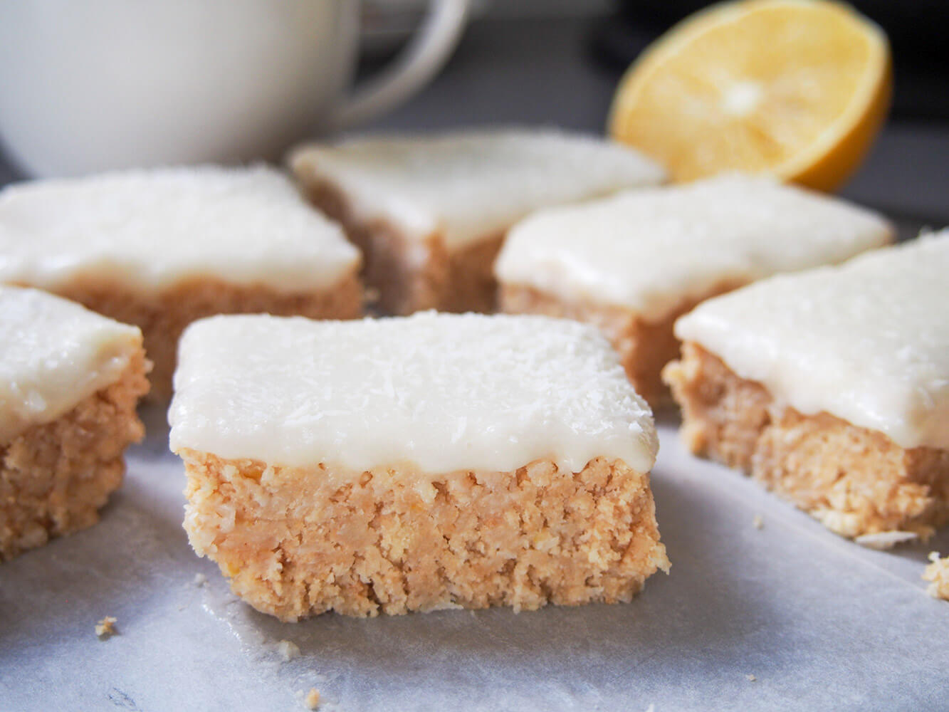 This lemon slice is a traditional Australian no bake bar with a coconut-cookie base and lemon frosting. It's really easy to make, with a wonderful sweet-tart flavor and perfect for so many occasions. lemon slice pieces with part lemon and coffee mug behind