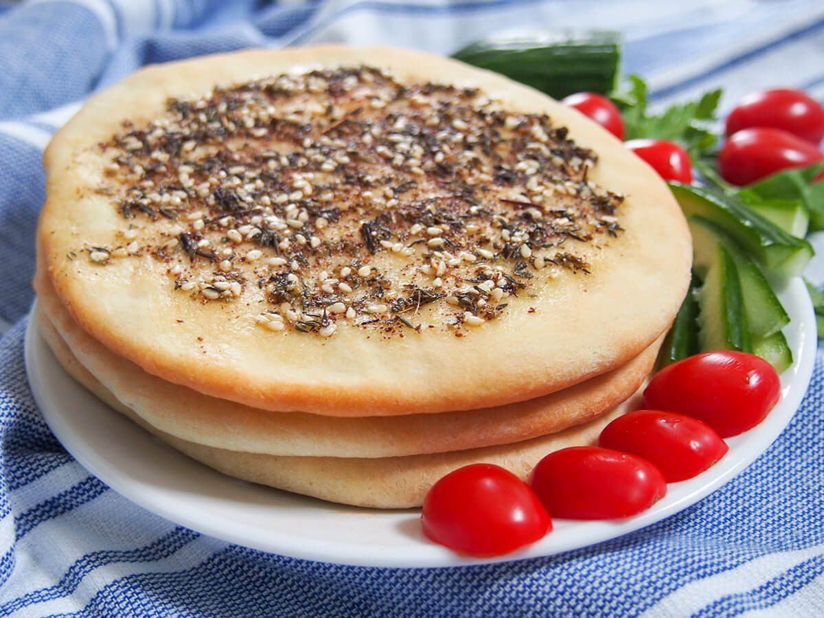 Manakish za'atar are delicious flatbread from the Levantine region topped with olive oil and za'atar. The oil and herbs add lots of aromatic flavor, making this bread perfect to snack on. stack of Manakish Lebanese za'atar flatbread with tomatoes and cucumber to side of plate