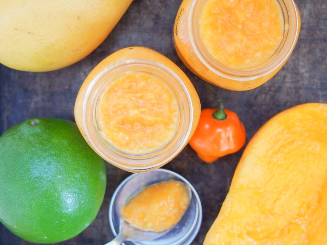 Mango habanero sauce gives a wonderful sweet-spicy kick to tacos and more. Really easy to make, and surprisingly healthy, it'll be a go-to hot sauce. two jars of mango habanero sauce from above with spoon of sauce resting on lid below and lime, chili and mango to side