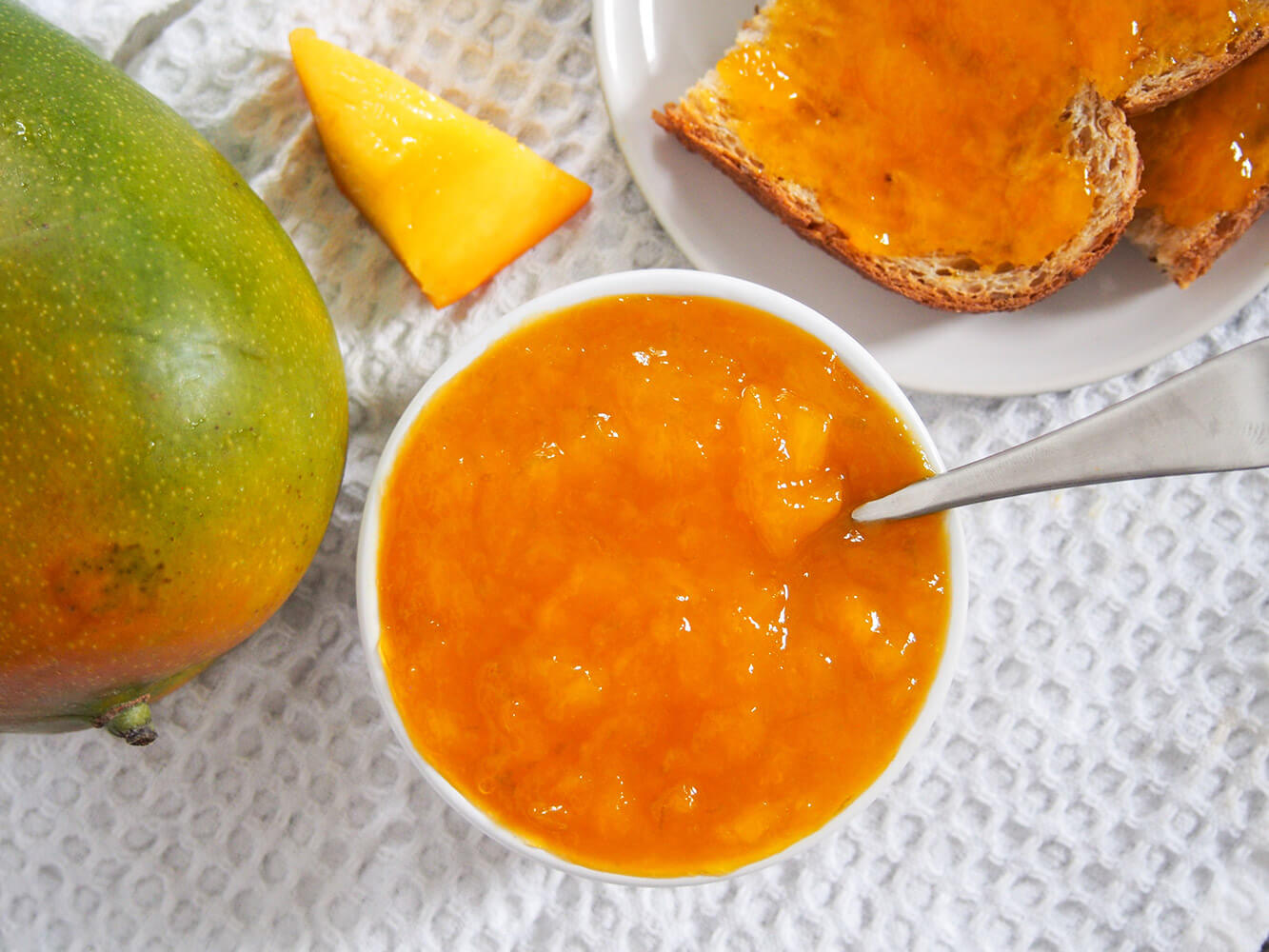 Enjoy some wonderful tropical flavors on your toast with this easy mango jam. It is quick to make, has a wonderful sweet flavor and is the perfect way to enjoy mango season that bit longer. bowl of mango jam with spoon in it with jam on toast to one side and mango to other