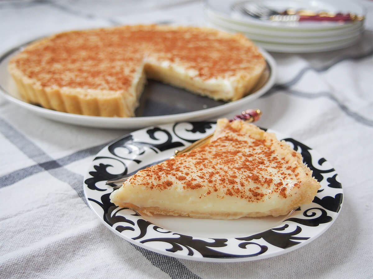 South African milk tart (melktert) slice of South African milk tart (melktert) in front with rest of tart on plate behind.