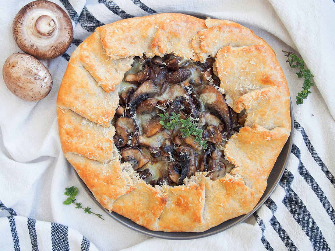 This mushroom galette is a delicious savory tart, with some garlic, thyme and fontina cheese adding flavor to the earthy mushroom filling. It makes a great light lunch, or bulk it out for a meatless meal. mushroom galette viewed from overhead with fresh mushrooms to side