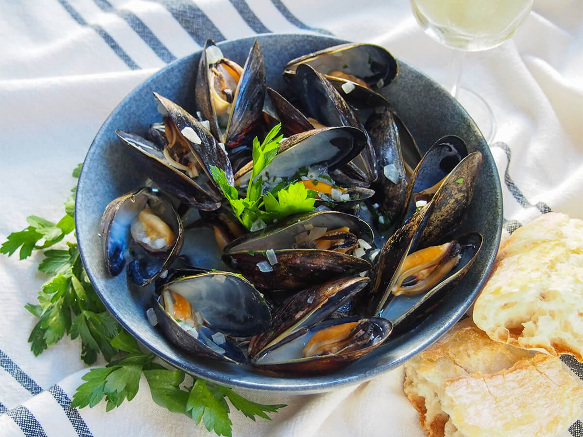Mussels in white wine is an incredibly simple and easy dish to make, but so delicious. Quick, tasty and one you'll want to make again soon. bowl of mussels in white wine with parsley around bowl and bread to one side