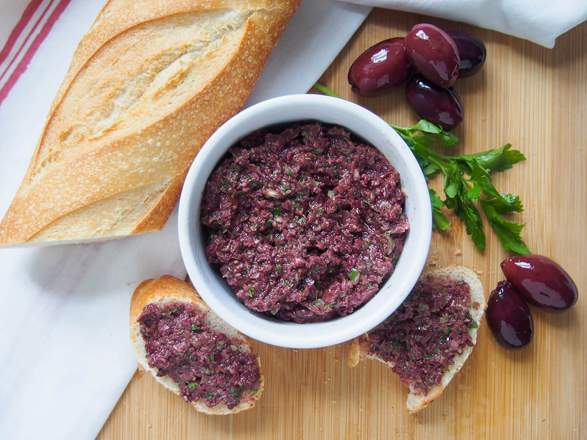 Tapenade is a delicious spread/sauce from Southern France made with olives, capers & anchovies. It's easy to make and so versatile - try it on bread, in pasta and more. dish of tapenade with part of baguette to side and olives and parsley to other