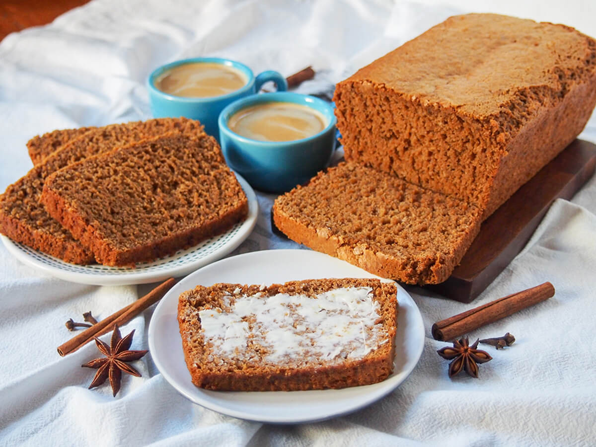 ontbijtkoek is a lovely Dutch spiced cake or breakfast cake buttered slice of ontbijtkoek Dutch spice cake with loaf and more slices behind and coffee cups in middle back.