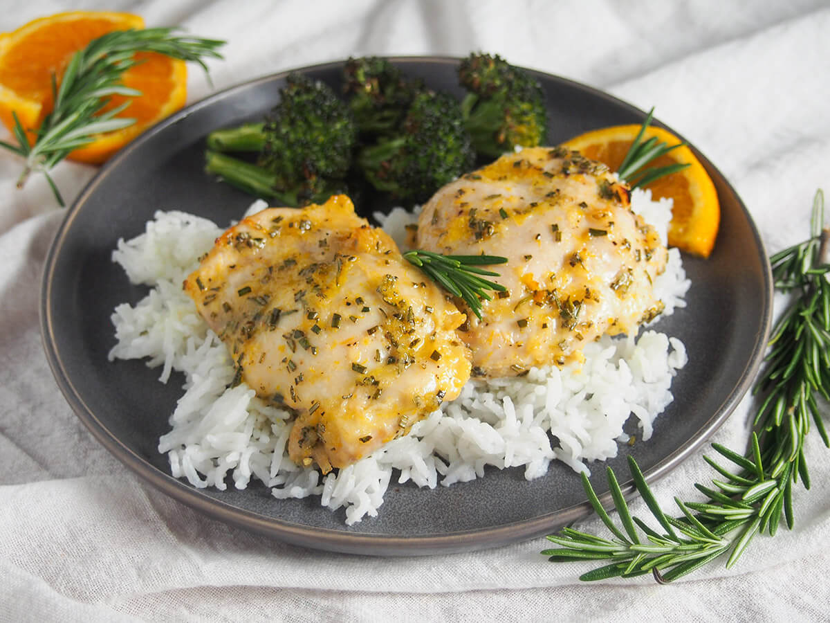 This orange rosemary chicken has a lovely combination of gently sweet and herb flavors. It's easy to prepare and perfect for any night of the week. The simple marinade comes together in no time, and then makes a tasty sauce for serving. plate with two pieces of orange rosemary chicken over rice with broccoli behind and sprig of rosemary in front of plate