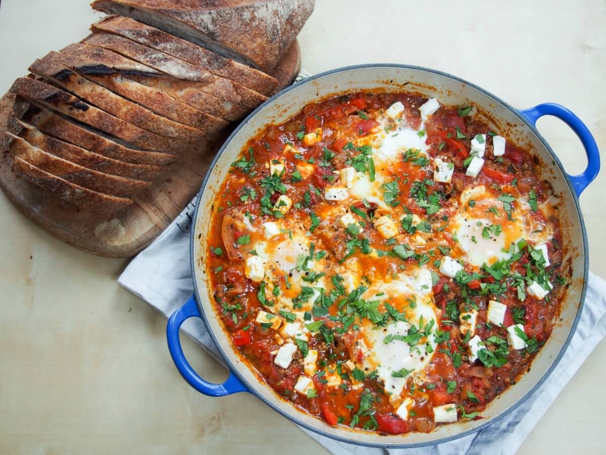beefed-up shakshuka (shakshuka with meat) - a delicious, hearty dish with a tomato, pepper and onion base, lots of herbs and topped with eggs and feta. Makes a delicious brunch/lunch or enjoy it for 'brinner'! shakshuka with meat in dish with sliced loaf of bread to side
