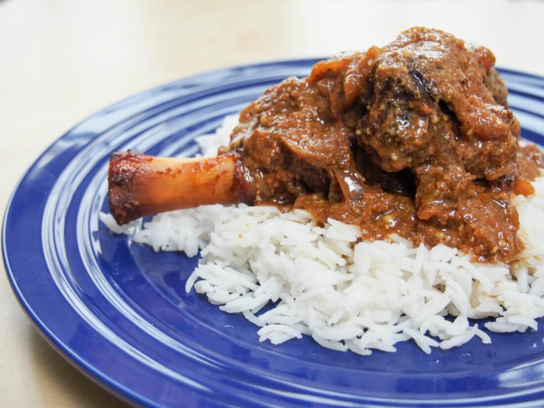 This delicious lamb rogan josh is made with lamb shanks in the slow cooker. Easy, meltingly tender & incredibly delicious: so full of aromatic Indian flavor lamb shank with rogan josh sauce over rice on dark blue plate