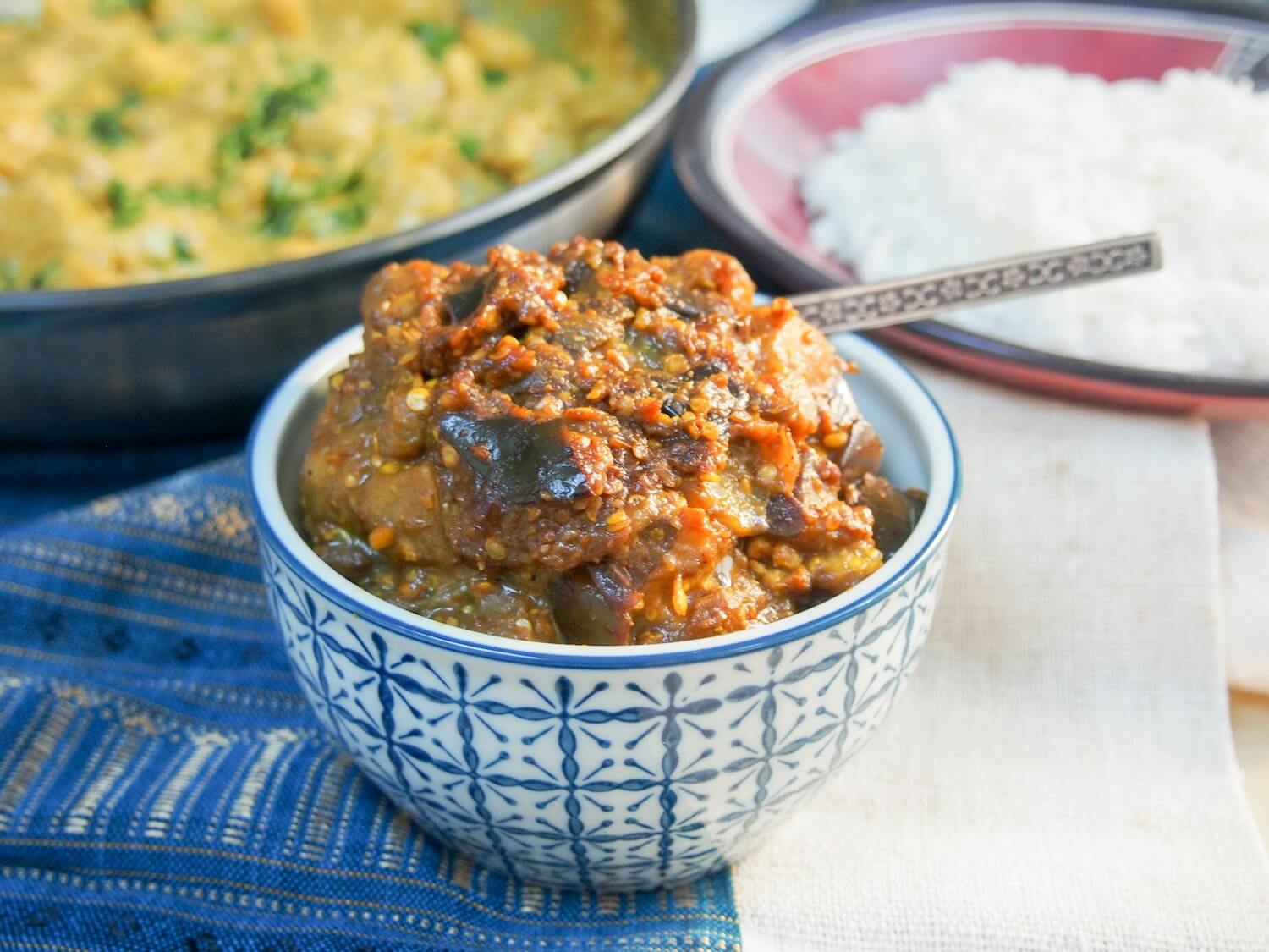 Brinjal pickle (eggplant relish/aubergine chutney) is a classic Indian condiment that is the perfect addition to any curry. Slightly sweet and spicy, it's easy to make. brinjal pickle (Indian eggplant relish/aubergine chutney) with spoon in dish and plates of curry and rice behind
