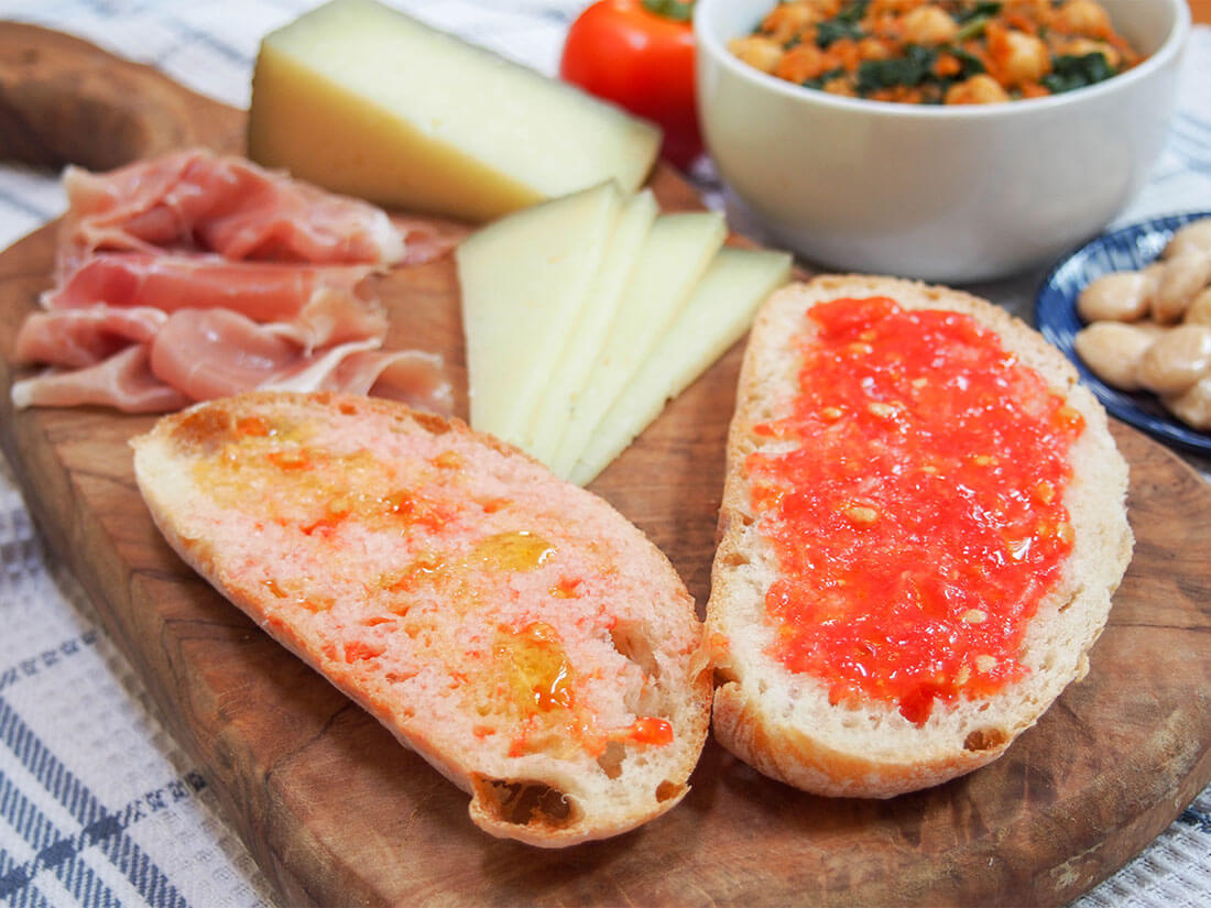 Pan con tomate - "bread with tomato" - is a Spanish tapas classic. It's a super easy combination but it transforms plain old bread into a tasty side or base for an open sandwich. Despite being so easy, there's more than one way to make it , and both are delicious! Pan con tomate (pa amb tomaquet) on cheese board with slices of ham and cheese behind