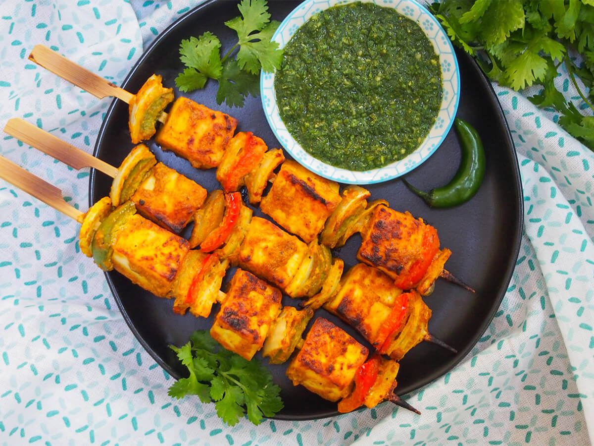 Paneer tikka are a bright and tasty vegetarian appetizer or snack, combining chunks of mild cheese and vegetables with a spiced marinade. They're really easy to make and perfect for entertaining or any excuse. three paneer tikka skewers on plate with green chutney above