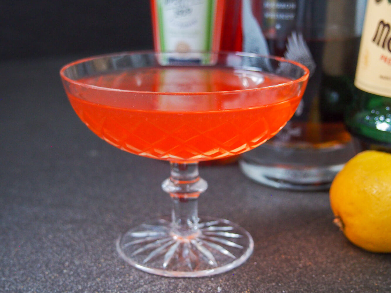 The paper plane cocktail is considered a modern classic by some. It's elegant, well balanced and easy to make, with the perfect balance of sweet, tart and spirit-forward flavor. Perfect for aperitivo hour. glass of paper plane cocktail with bottles behind and lemon to side
