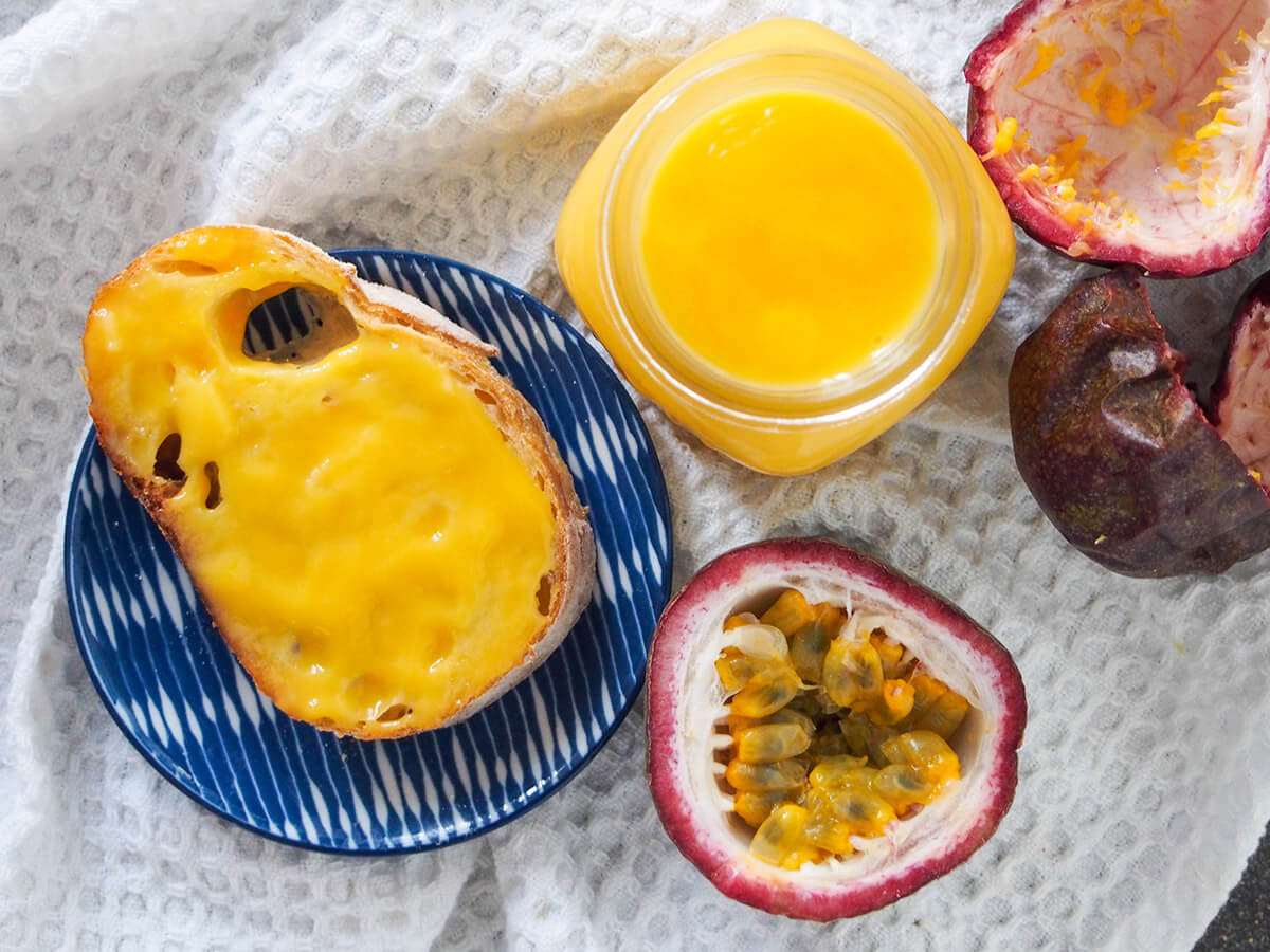 This passion fruit curd is a tropical twist on the classic citrus spread. It’s bright and aromatic, with that classic smooth texture. Easy and delicious! jar of passion fruit curd with bread with curd to side and passion fruit below, empty passion fruit shell to other side