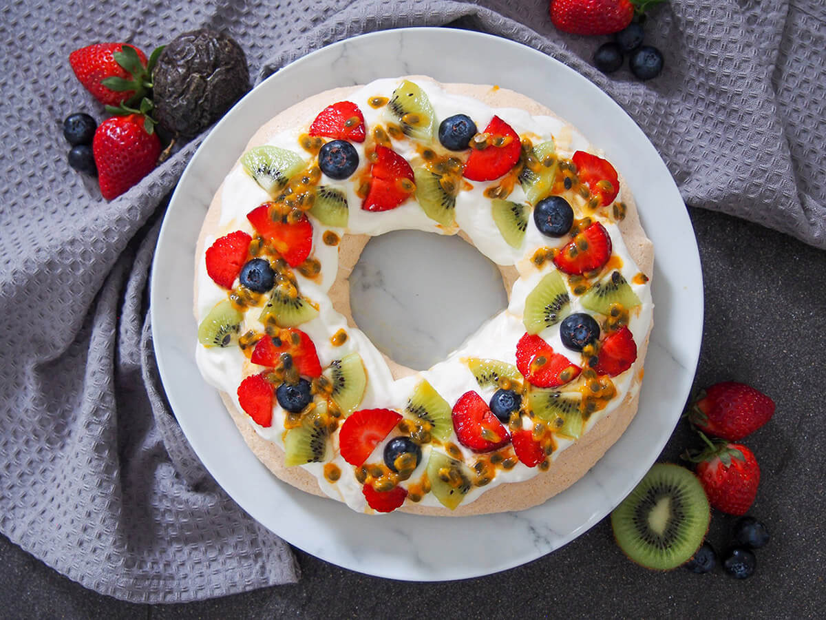 This pavlova wreath is a festive twist on a classic dessert. The combination of meringue with a chewy middle, cream and fruit is delicious & elegant. pavlova wreath topped with cream and fruit with additional fruit to side of plate