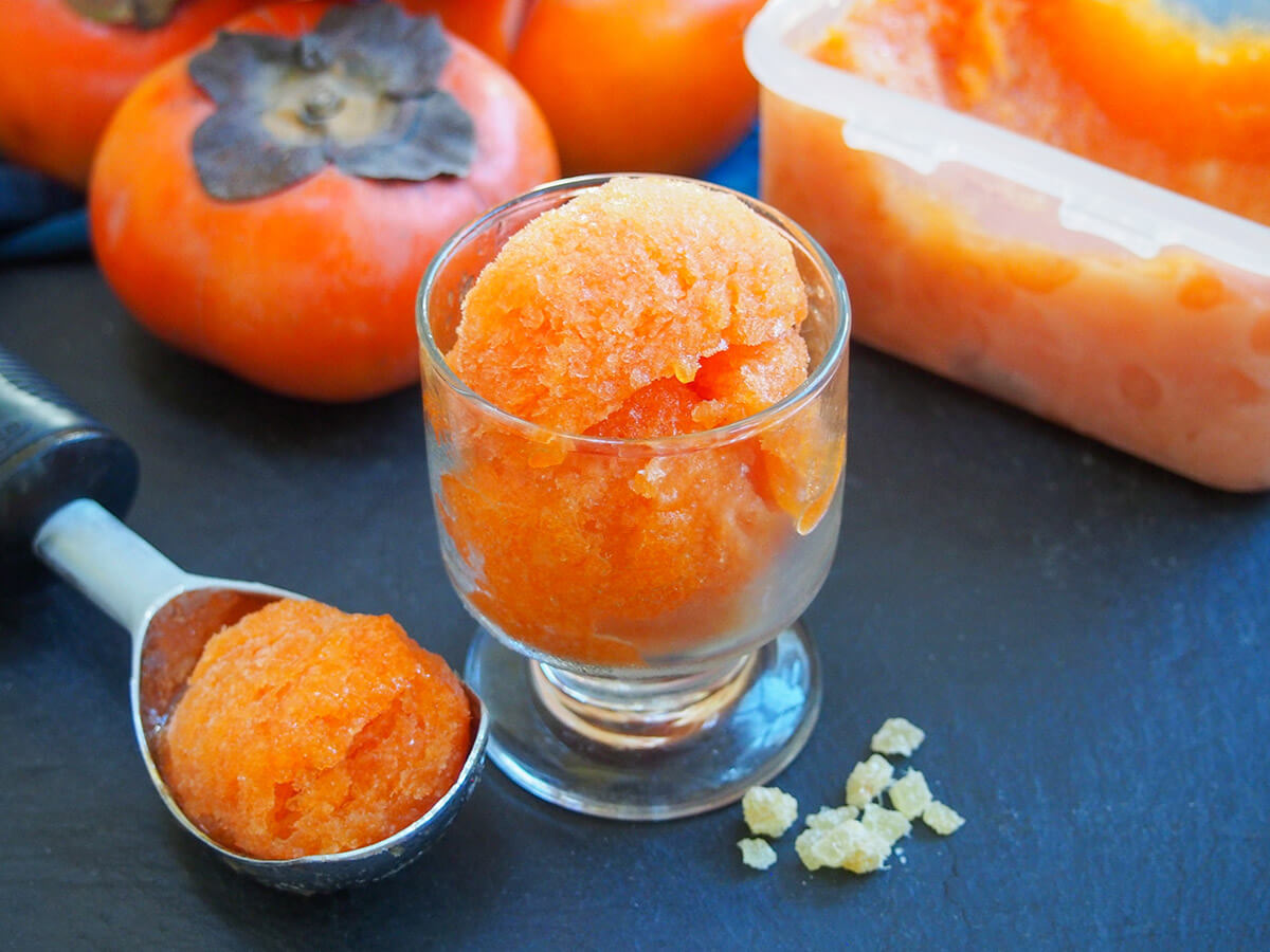 This delicious persimmon ginger sorbet is easy to make and both looks and tastes great. The zingy ginger compliments the smooth, fragrant persimmon perfectly. glass with persimmon ginger sorbet with scoop to side and whole fruit behind