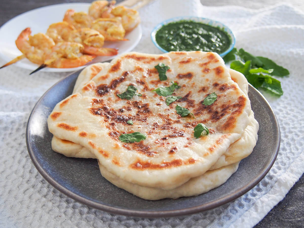 Peshwari naan is a wonderfully tasty filled bread that combines a gently sweet, nutty filling with soft and chewy dough. It's a classic accompaniment to Indian meal that's easy to recreate at home to go with a meal or enjoy as a snack. peshwari naan on plate with shrimp and chutney behind