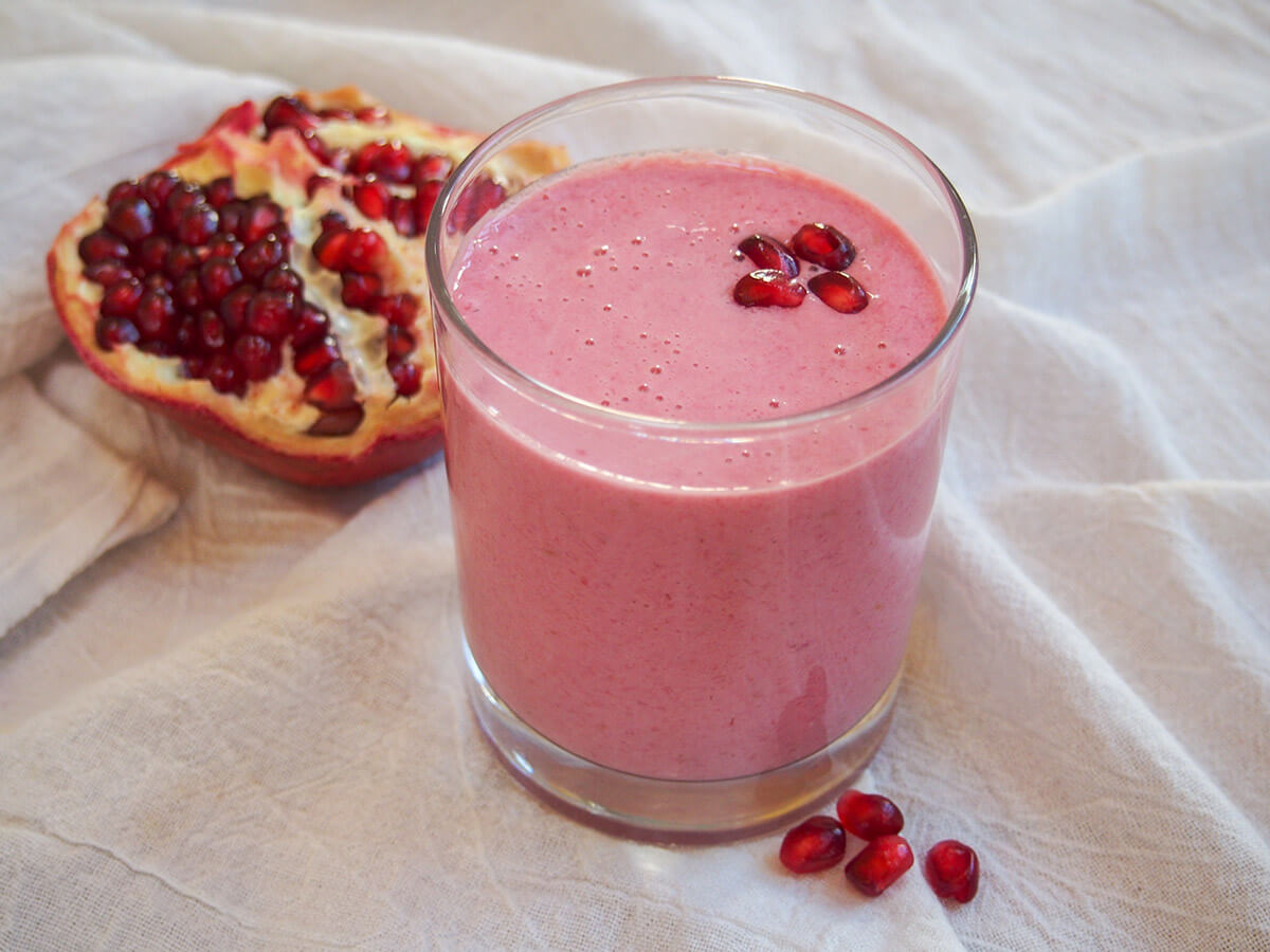 This pomegranate smoothie is super easy to make and has a tasty balance of bright and sweet berries, sweet-tart pomegranate and smooth yogurt. Perfect as a pick me up or easy breakfast. glass of pomegranate smoothie with part of pomegranate behind