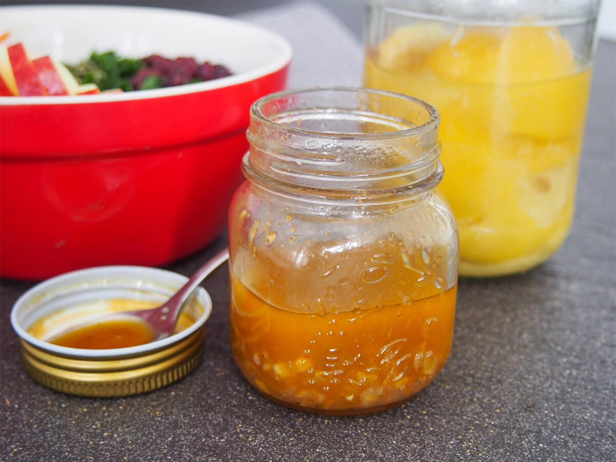 This preserved lemon vinaigrette/dressing is a delicious mix of tangy lemon and warm spice flavors. It works particularly well with crunchy autumnal/winter salads but would also be great mixed through couscous and more. Jar of preserved lemon vinaigrette in front of bowl of salad and jar of preserved lemons