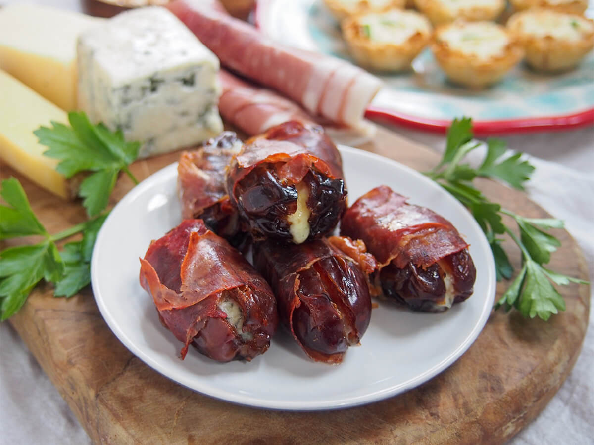 These prosciutto wrapped dates have only 3 ingredients, take no time to prepare and are a wonderful sweet-salty mix. They're addictively tasty little bites, perfect for any occasion. plate of prosciutto wrapped dates on wooden board with cheese and ham behind