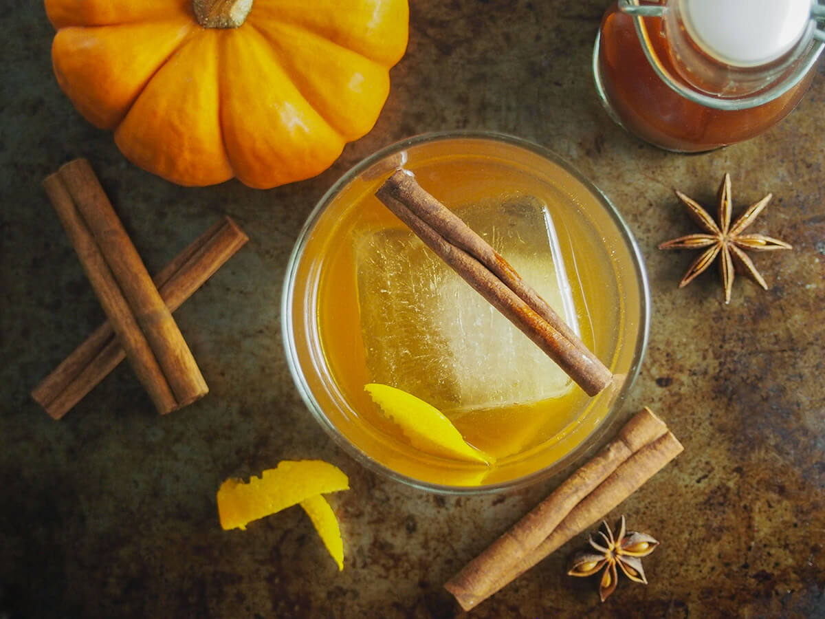 pumpkin spice old fashioned is a tasty twist on the classic cocktail. glass of pumpkin spice old fashioned with whole spices, mini pumpkin and bottle of spiced syrup around