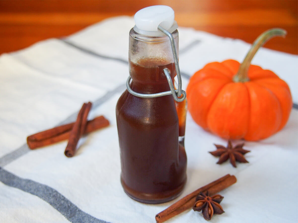 Homemade pumpkin spice syrup is easy to make and adds a wonderful warm sweet spice flavor to coffee, cocktails and more. bottle of homemade pumpkin spice syrup with spices to side and mini pumpkin behind