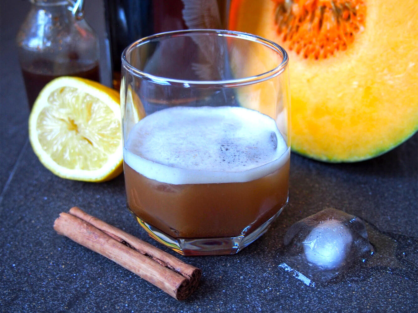 This pumpkin spice whiskey sour is the pumpkin spice cocktail you've been searching for! It's easy to make, with a wonderful balance of warm spices and tart lemon. A wonderful warm twist on a classic. glass of pumpkin spice whiskey sour with cinnamon stick, lemon and pumpkin around glass