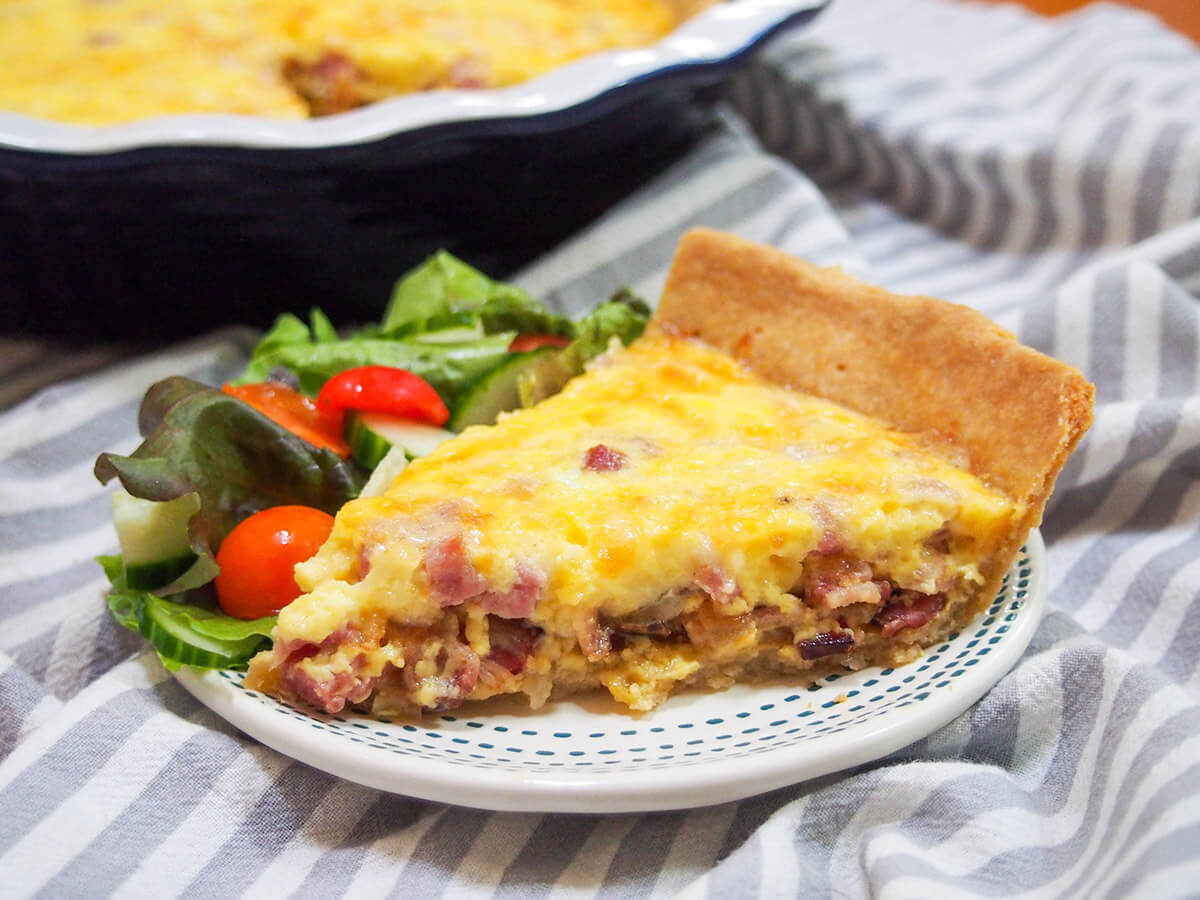 Quiche Lorraine is a classic French savory tart packed with gruyere cheese, bacon and smoked ham. Comforting and delicious, it makes a great lunch. slice of quiche Lorraine with salad on plate