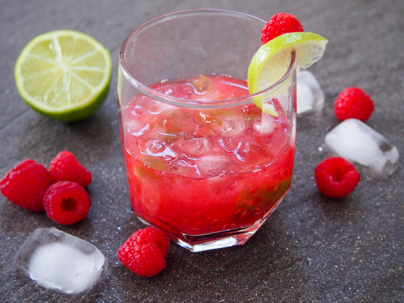 This raspberry caipirinha is a bright and fruity twist on the classic Brazilian cocktail. It's quick and easy to make, and perfect to sip on a warm day. raspberry caipirinha in glass with raspberries and lime to side