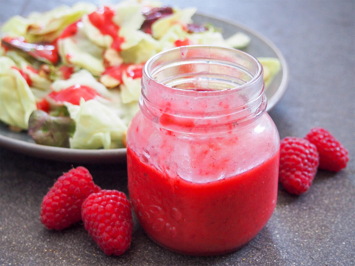 This raspberry vinaigrette is as bright in flavor as its color. It comes together easily and adds a wonderful burst of flavor such as to simple salads for a quick and easy side. Plus this one is on the healthier side, too. jar of raspberry vinaigrette with raspberries to side