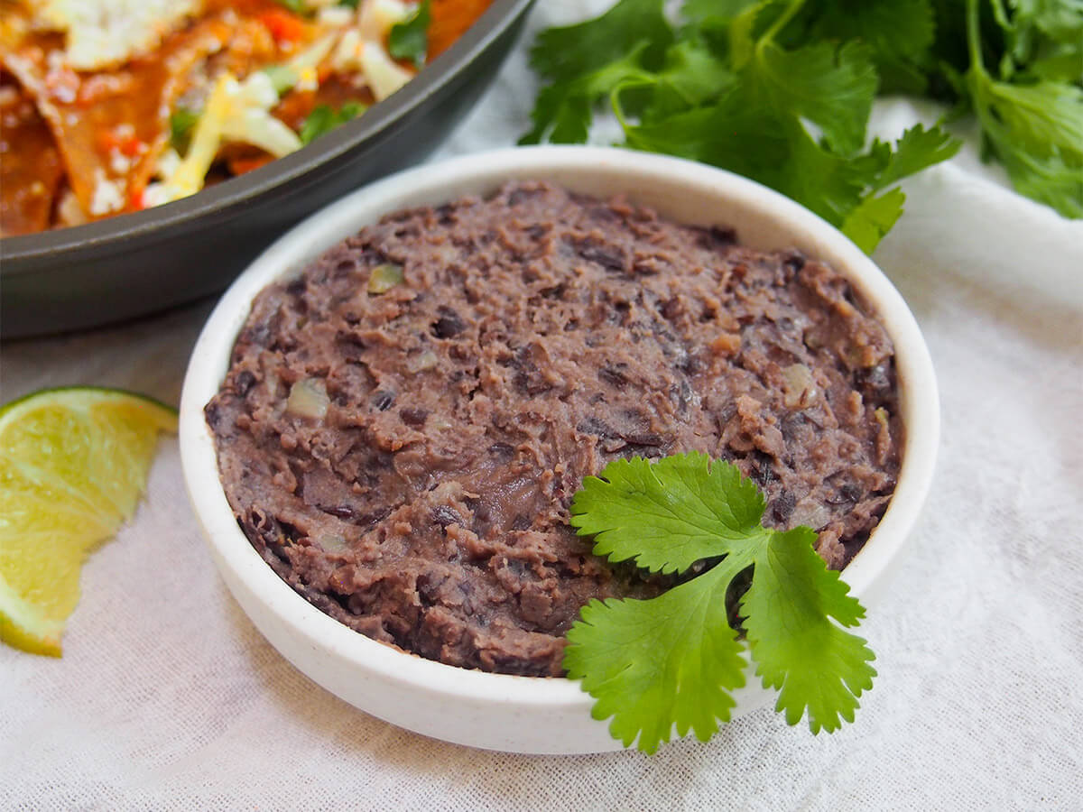 Homemade refried black beans are so much easier to make than you might think. You only need a handful of ingredients and a couple minutes for a tasty, versatile side that's perfect for Mexican sandwiches, breakfasts, tostadas and more. dish of refried black beans with cilantro and other food part in view behind