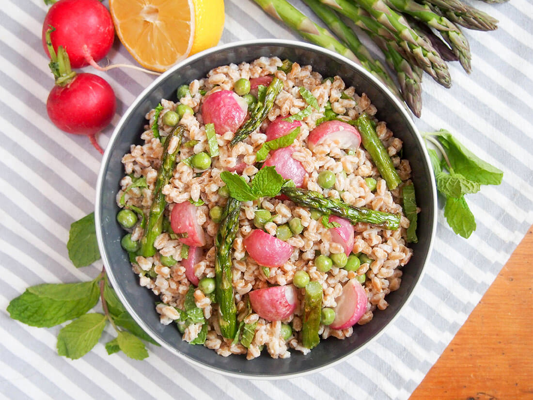 This roasted spring vegetable farro salad is a delicious and versatile way to enjoy the flavors of spring. Pack it for lunch or eat it as a side. The mellow flavors and heartiness of the farro are just what a spring day needs! Roasted spring vegetable farro salad in bowl viewed from overhead with ingredients around bowl