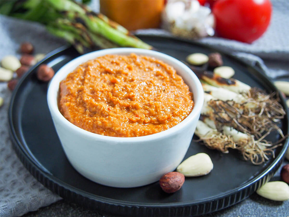Romesco sauce is a delicious, versatile Spanish sauce made with tomatoes, almonds, hazelnuts and peppers. Made the traditional way takes a little more time, but it's still easy and worth it for the wonderful depth of flavor. Great with grilled vegetables, fish, meat and more. romesco sauce in dish with grilled spring onions on plate behind
