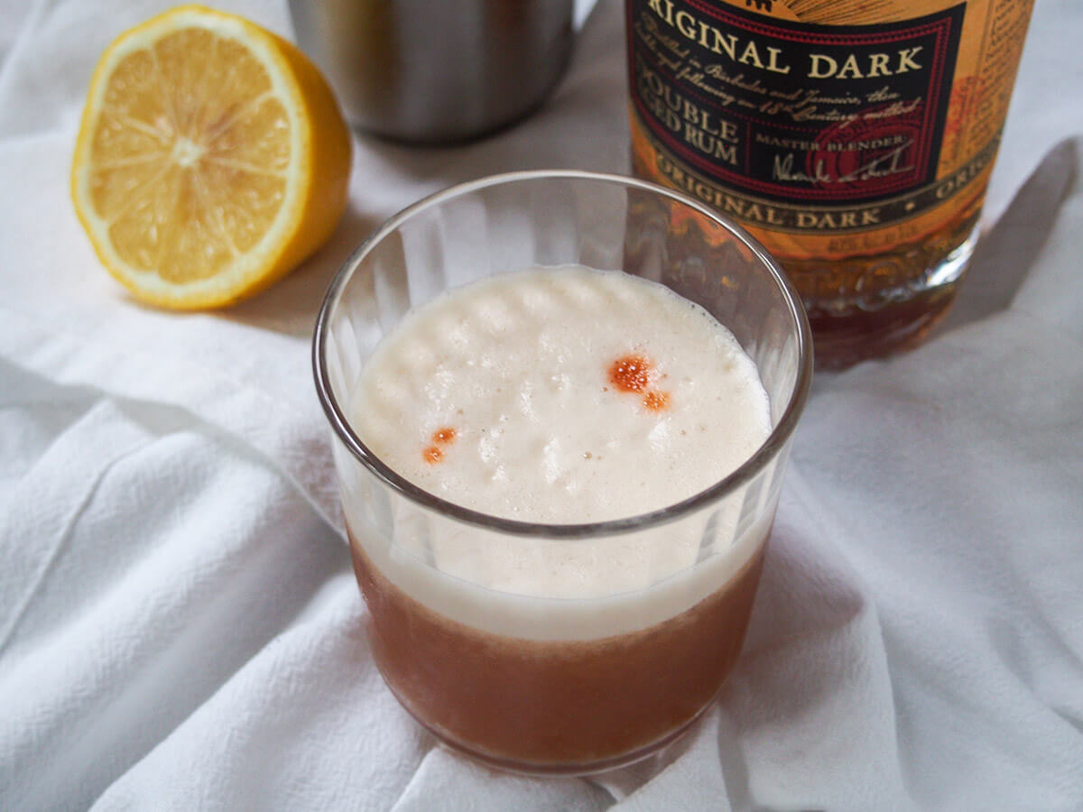 This rum sour is a tasty take on the sour cocktail with depth from dark rum and a lovely tart balance. Easy to make and great for sipping. glass of rum sour with rum bottle and half lemon behind.