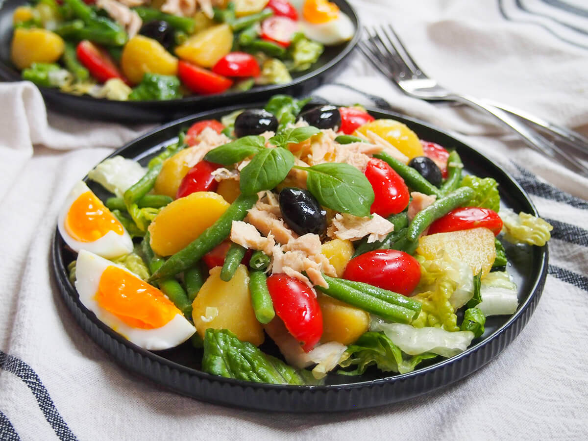 This bright and varied Nicoise salad combines fresh vegetables, potatoes, beans and fish. A delicious combination, enough to make a meal or divide further for a tasty appetizer. plate of salade Nicoise garnished with basil