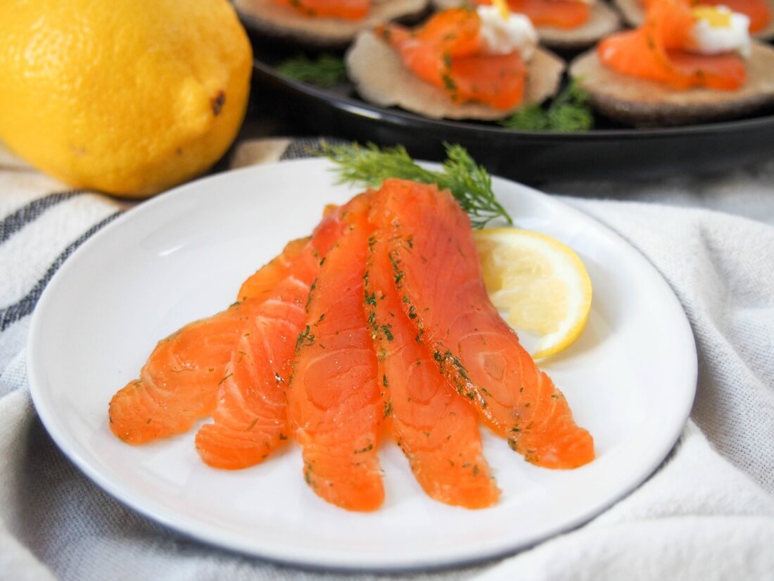 Salmon gravlax tastes similar to smoked salmon, but without the pricetag. Easy to make, all you need is a little planning, then enjoy the delicate, lightly herbed flavor of this home-cured salmon. Great as an appetizer and more. salmon gravlax on plate with a lemon and some gravlax topped blini behind