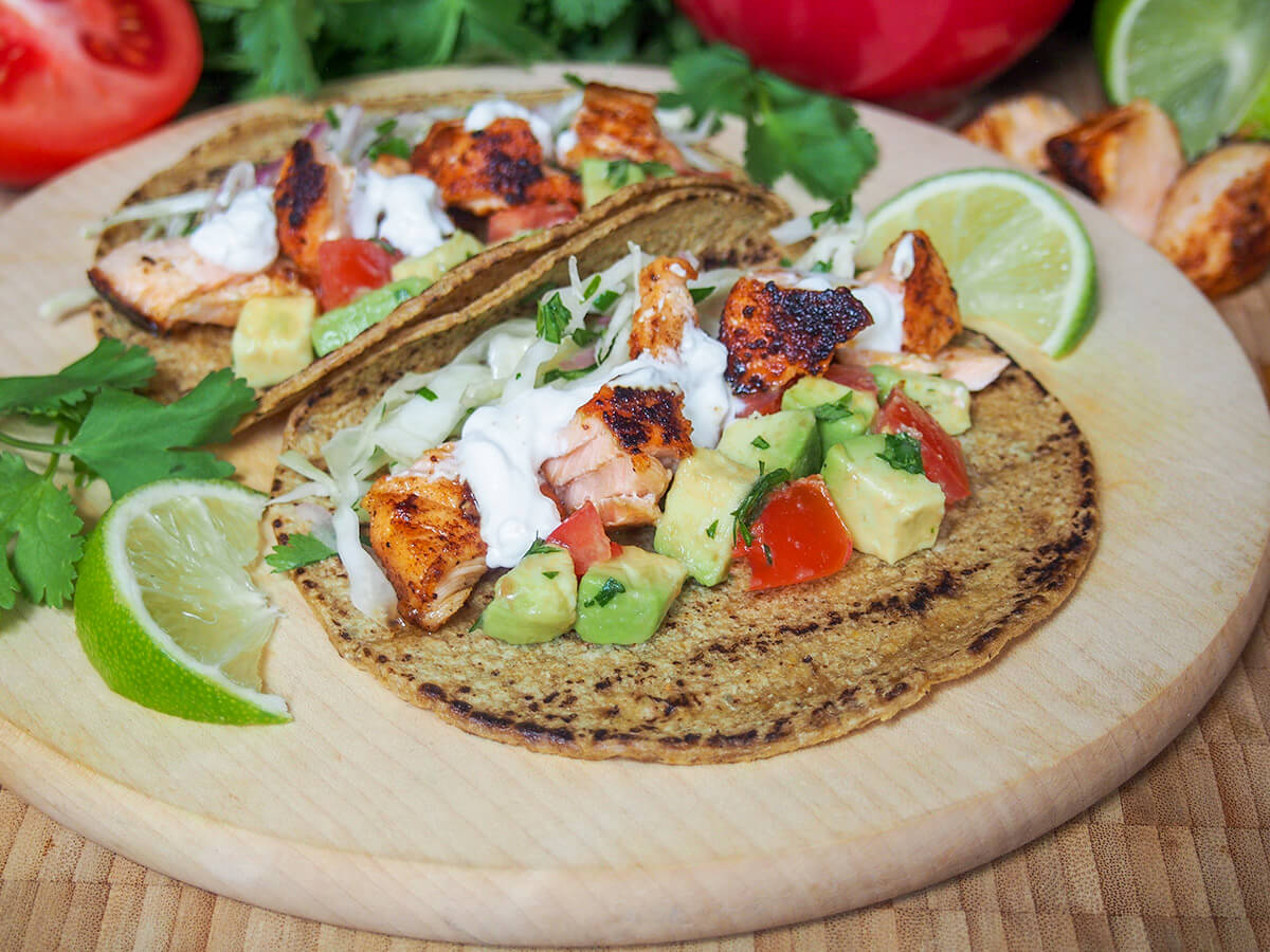These salmon tacos are easy, healthy & delicious. Served with a simple cilantro cabbage slaw and avocado-tomato salsa, it's a tasty mix of flavors and textures. salmon taco with another sitting behind it on a wooden board with slices of lime to either side