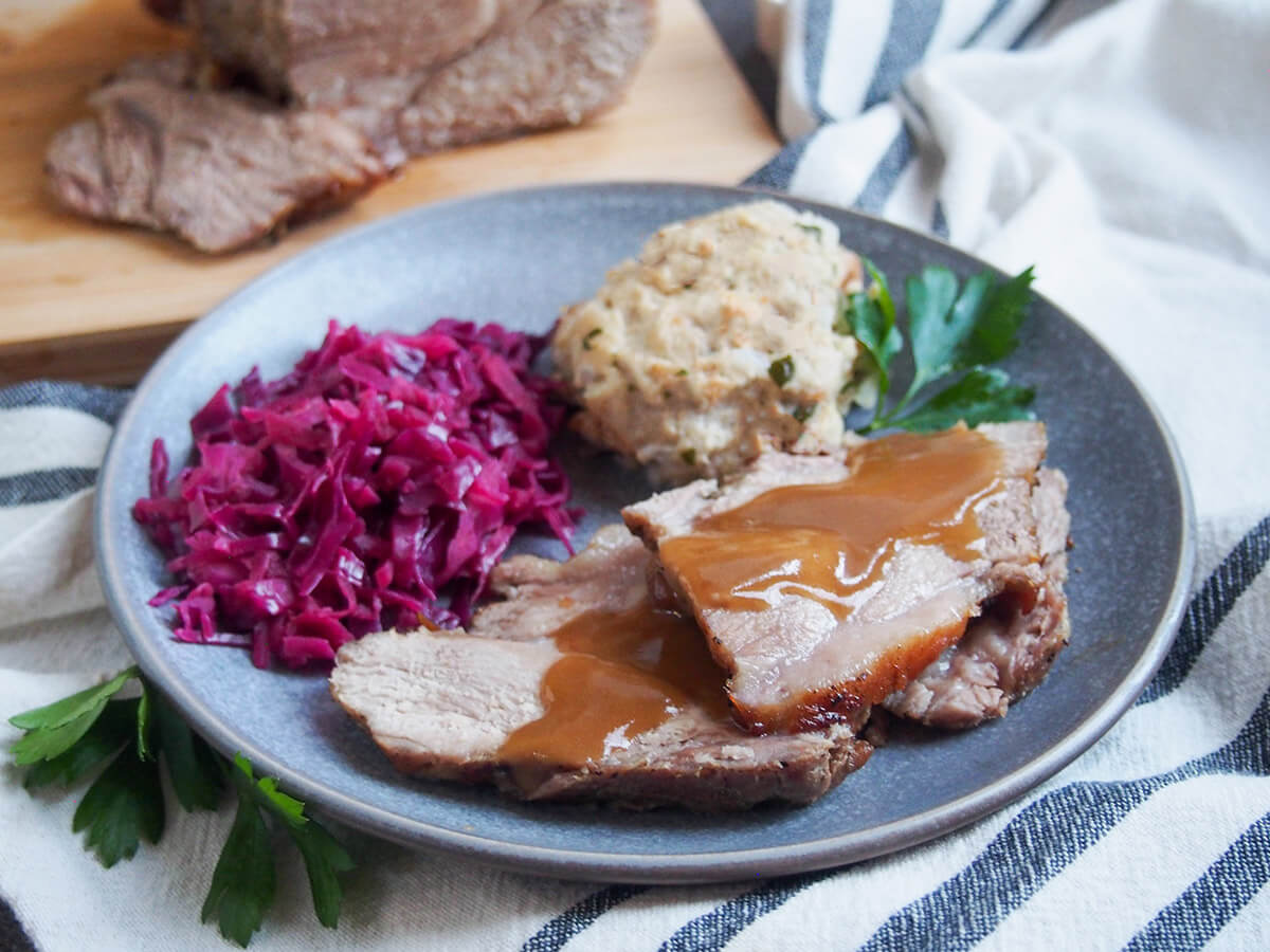 Schweinebraten is a traditional German pork roast, and in many cases, as here, it's cooked with beer which helps make a delicious sauce. The seasonings are simple but the result is a deliciously tender, flavorful main. slices of Schweinebraten - German pork roast - on plate with red cabbage and dumpling behind