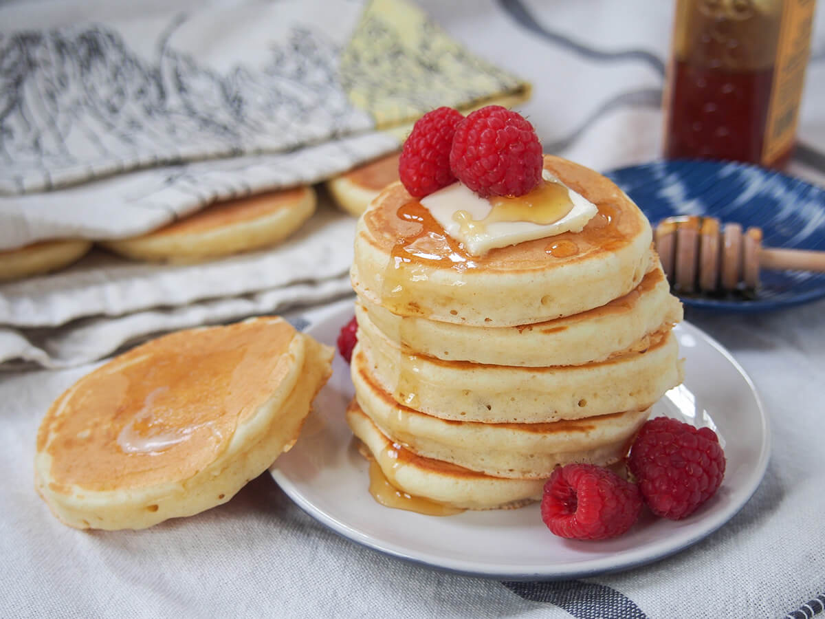 Scotch pancakes stack of Scotch pancakes on plate with raspberries to side and another pancake resting on side of plate with butter melted on top.