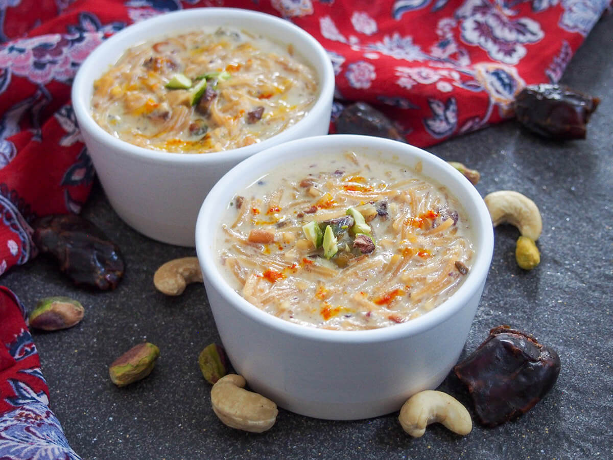 Sheer khurma is a dessert made from milk simmered with dates and other dried fruits, and loaded with nuts and vermicelli. It's creamy and comforting, and a classic dish for Eid-al-Fitr. bowl of sheer khurma with a second bowl behind and nuts and dates around bowls