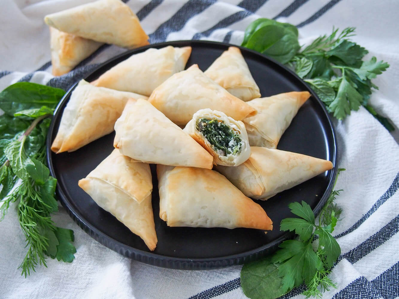 Spanakopita triangles are a great finger-food version of the classic Greek spinach and feta phyllo pie. They're crisp and tasty, and much easier to make than you might think. spanakopita triangles on plate with one bitten into, resting on others showing filling inside