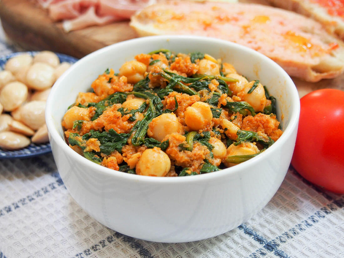 Spanish chickpeas and spinach are an easy and delicious vegetarian tapas dish Spanish chickpeas and spinach in bowl with pan con tomate and almonds behind