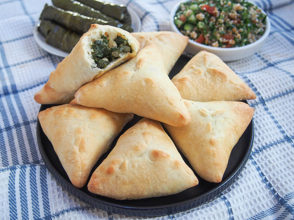 Spinach fatayer are a kind spinach hand pie with simple ingredients yet plenty of flavor. Perfect as part of a mezze meal, as a snack or any excuse you like. plate of spinach fatayer with plates of grape leaves and tabbouleh behind