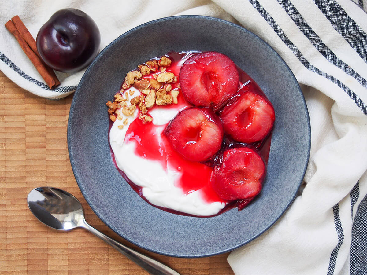 Stewed plums are a really quick and easy way to prepare plums in a sweet syrup with a hint of spice. Perfect for breakfast with yogurt or as dessert with ice cream. Tender and delicious. bowl of stewed plums over yogurt with spoon below bowl and plum and cinnamon stick to side