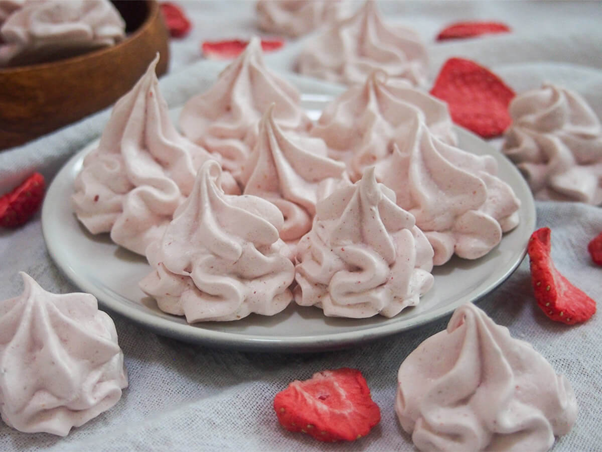 These strawberry meringue cookies are easy to make, with a wonderful delicate color, crunch and flavor. Perfect to snack on, decorate other desserts & more. plate of strawberry meringue cookies with dried strawberries around plate