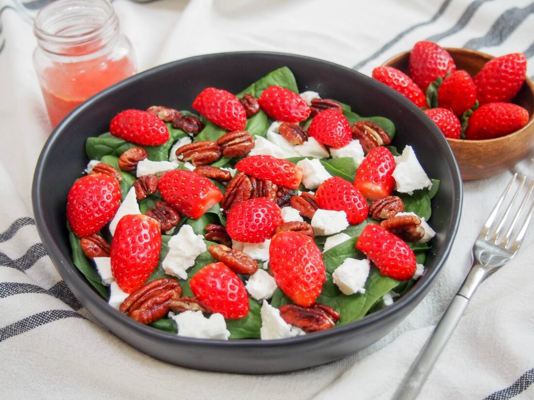 This strawberry spinach feta salad is a simple and delicious way to eat healthily, without skimping on flavor. With bright colors, a lovely mix of fresh flavors and crunch, it's perfect for lunch or as a side. bowl of strawberry spinach feta salad with strawberries to side, dressing in jar behind and fork to side of bowl