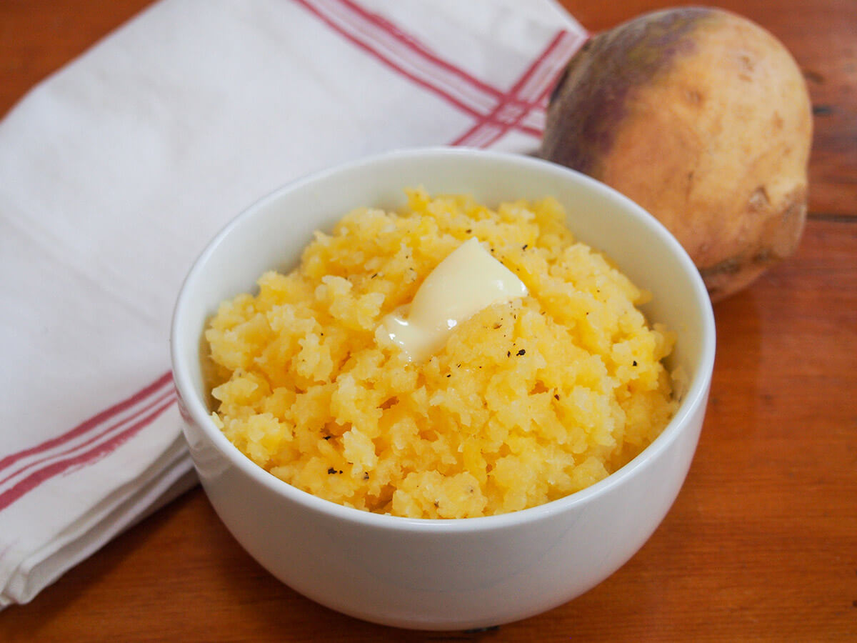 Mashed rutabaga, or swede mash, is an easy side dish and tasty alternative to mashed potato or other root vegetables. It pairs perfectly with a range of mains like roast meats and sausages with a mild and gently sweet flavor. bowl of mashed rutabaga with cloth and rutabaga/swede behind