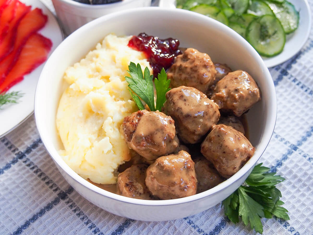 Forget the frozen version, traditional Swedish meatballs (köttbullar) are easy to make at home and taste delicious. These gently spiced meatballs in a flavorful cream sauce are classic comfort food. bowl of Swedish meatballs with mashed potato with plates of cucumber and gravlax in background