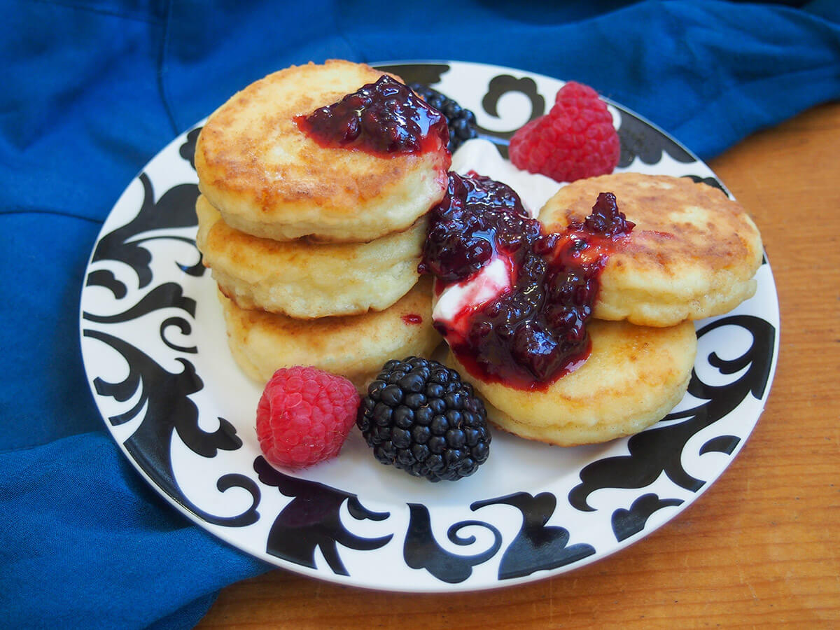 Syrniki are small, thick Ukrainian/Russian sweet cheese pancakes. They might not have a lot of ingredients and look a bit unassuming, but these little bites are soft, fluffy and deliciously good. plate of syrniki Russian cheese pancakes with jam, sour cream and fruit